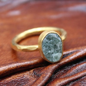May include: A gold-colored ring with a textured band and a bezel-set, oval-shaped, green-grey stone. The ring is displayed on a brown, textured surface, showcasing the jewelry's design and color.