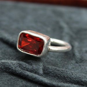 May include: A silver ring featuring a rectangular, deep red gemstone. The gemstone is set in a silver bezel, and the band is a simple, rounded design. The ring is displayed on a dark, textured surface.