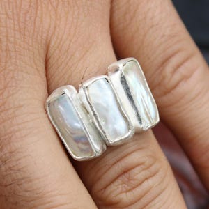 May include: A silver ring featuring three rectangular, iridescent, white pearl stones. The ring is set in a polished silver band. The ring is shown on a person's finger.