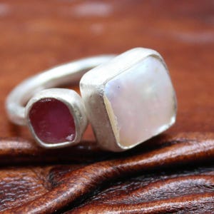 May include: A silver ring featuring two gemstones. One is a square, iridescent, off-white stone, and the other is a smaller, oval-shaped, deep pink stone. The ring is set against a brown leather surface.