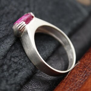 May include: A silver ring with a large, oval-cut, deep pink gemstone. The ring has a simple, modern design with a brushed metal finish. The gemstone is set in a bezel setting, and the ring is displayed on a dark, textured surface.