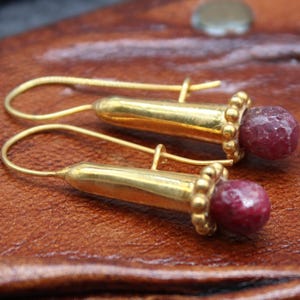 May include: A pair of gold-colored earrings with a hook design. Each earring features a cylindrical gold element with a beaded base, topped with a faceted, deep red gemstone. The earrings rest on a brown leather surface.