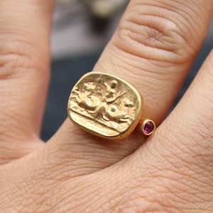 May include: A gold ring featuring a rectangular face with a detailed relief design. A small, round, red gemstone is set on the band. The ring is worn on a finger, showcasing the intricate craftsmanship and design.