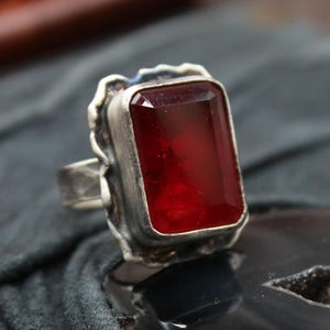 Puede incluir: Un anillo de plata con una gran gema rectangular de color rojo intenso. La gema está engastada en un bisel de plata texturizado y ornamentado. La banda del anillo es de plata, con un diseño sencillo. El estilo general es vintage o antiguo.