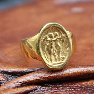 Può includere: Un anello dorato con una faccia ovale con un rilievo di due figure. L'anello è posto su una superficie marrone strutturata. Il design dell'anello suggerisce un tema classico o storico.