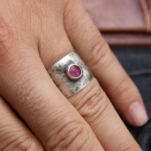 May include: A wide, hammered silver ring with a round, bright pink gemstone set in the center. The ring is worn on a finger, showcasing the textured metal and the vibrant color of the stone. The ring is a piece of jewelry.