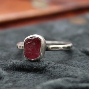 May include: A silver ring with a rectangular, deep red gemstone. The band has a hammered texture, and the stone is set in a smooth bezel. The ring is displayed on a dark gray surface.