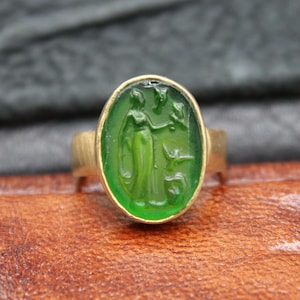 Può includere: Un anello d'oro con una gemma verde ovale. La gemma presenta un disegno intagliato di una figura e altri elementi. L'anello è posto su una superficie di cuoio marrone.
