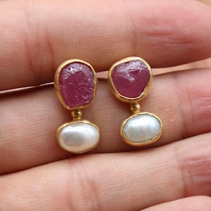 May include: A pair of gold-toned earrings featuring a ruby gemstone at the top and a white pearl at the bottom. The earrings have a unique design with a small gold connector between the two stones. The earrings are set against a skin-toned background.