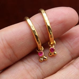 May include: A pair of gold-colored hoop earrings with a hammered texture. Each earring features a small, dangling charm with a single, round, red gemstone. The earrings are displayed against a blurred, brown background.