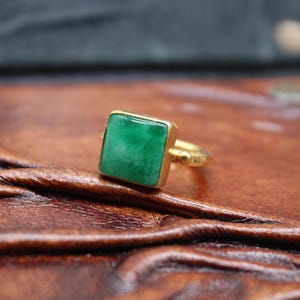 May include: A gold ring with a square, vibrant green jade stone. The ring's band has a textured design. The ring is displayed on a textured, brown leather surface, with a dark background.