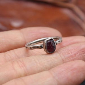 May include: A silver ring with a dark red gemstone. The ring has a hammered texture on the band and a bezel setting for the stone. The gemstone is a deep, rich red color and is the focal point of the ring.