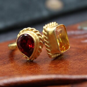 Könnte beinhalten: Zwei goldene Ringe mit Edelsteinen. Ein Ring hat einen birnenförmigen, tiefroten Edelstein. Der andere Ring hat einen quadratischen, gelben Edelstein. Beide Ringe haben ein gedrehtes Seildesign um die Edelsteine.