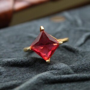 May include: A gold ring featuring a large, square-cut, vibrant red gemstone. The ring's band is a simple, thin design, and the gemstone is set with four prongs. The ring is displayed on a dark gray, textured surface.