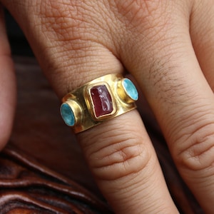 May include: A gold-colored ring with a rectangular red gemstone in the center and two round turquoise gemstones on either side. The ring is worn on a finger, showcasing the jewelry's design and color contrast.