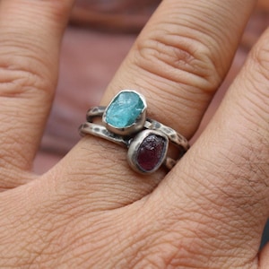 May include: Two stacked rings with raw gemstone settings. The top ring features a light blue stone, while the bottom ring showcases a deep red stone. The rings have a hammered silver band, creating a rustic, handcrafted aesthetic.