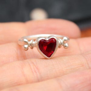May include: A silver ring with a heart-shaped, deep red gemstone. The ring features decorative accents on either side of the heart. The ring is held in a person's hand.