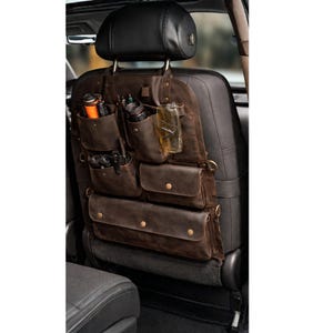 May include: A brown leather car seat organizer with multiple pockets and compartments. The organizer hangs from the headrest and features various sized pockets, including a drink holder. Several items are stored in the pockets, including pens, a flashlight, and a glass.