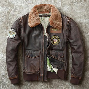 Puede incluir: Chaqueta bomber de cuero marrón con cuello de borrego. La chaqueta presenta varios parches, incluido uno que dice "USS Norfolk Naval Div." y otro que dice "Marine Service". Tiene dos bolsillos delanteros y un dobladillo y puños acanalados.