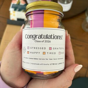 May include: A glass jar with a gold lid filled with colorful paper strips. The jar has a label that reads "Congratulations! Class of 2026" and lists emotions. The jar is held in a person's hands.