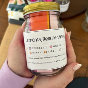 May include: A glass jar with a gold lid, filled with colorful paper strips. The jar has a label that reads "Grandma, Read Me When" and lists emotions. The jar contains 30 color-coded Bible verses. Handmade with love.