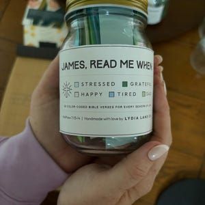 May include: A clear glass jar with a gold lid, labeled "JAMES, READ ME WHEN." The label includes checkboxes for emotions like "stressed" and "happy." The jar contains color-coded Bible verses. The text "Handmade with love by LYDIA LANE CO" is also visible.