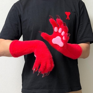 May include: A pair of bright red fuzzy gloves featuring long, dark, curved claws and light pink silicone paw pads on the palms. The gloves extend to the mid-forearm and are made of a soft, textured material that mimics animal fur.