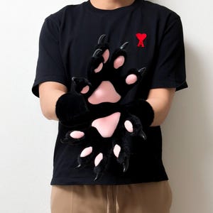 Könnte beinhalten: Ein Paar schwarze Plüschhandschuhe in Form von Tierpfoten mit weichen rosa Ballen und langen, gebogenen schwarzen Krallen. Die Handschuhe bestehen aus einem flauschigen Material und verfügen über eine detaillierte Polsterung an Handflächen und Fingern.