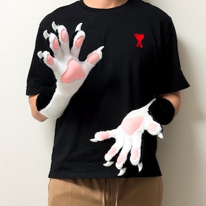 May include: A pair of plush animal paw gloves featuring white faux fur, black accents, soft pink paw pads, and pointed white claws. The gloves extend to the mid-forearm and are designed with five fingers, showcasing a detailed, textured appearance.