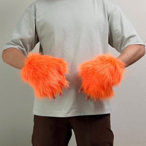 May include: A pair of bright orange faux fur fingerless gloves with a long, shaggy texture. The gloves cover the back of the hands and wrists, leaving the fingers exposed. The vibrant orange material has a dense, soft appearance.