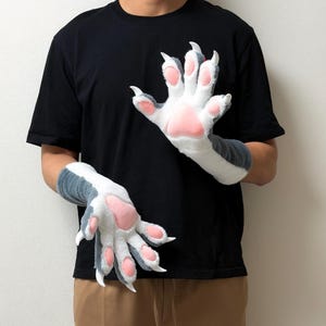 以下が含まれることがあります： 動物の足を模した、長いカフ付きのぬいぐるみのような手袋。白とダークグレーのフェイクファーを組み合わせ、手のひらと指には柔らかいピンク色の肉球が施され、指先には尖った白い爪が付いています。