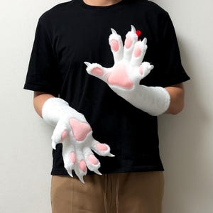 Könnte beinhalten: Ein Paar Handschuhe aus weißem Kunstfell in Form von Tierpfoten mit weichen, rosafarbenen Polstern und spitzen, weißen Krallen an jedem Finger. Die Handschuhe bedecken die Unterarme und bestehen aus einem dicken, flauschigen weißen Material.