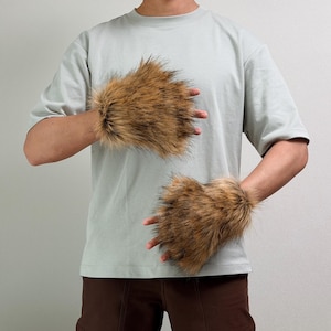 May include: A pair of fingerless gloves made from long, shaggy faux fur in shades of brown, tan, and black. The gloves cover the back of the hands and wrists, featuring a dense, textured appearance with varied dark and light hair tones.