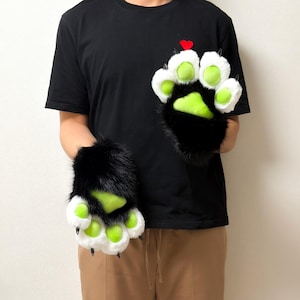 May include: A pair of plush animal paw gloves featuring black faux fur on the back, white fur around the fingers, and bright lime green paw pads. Each finger tip has a small black claw, and the gloves are worn over a black t-shirt.