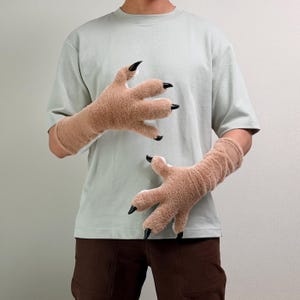 May include: A pair of tan, plush, long-cuffed gloves designed to resemble animal paws. The gloves feature a soft, fuzzy texture with four distinct finger sections, each tipped with a sharp, glossy black claw. The design covers the hands and forearms.