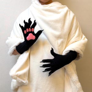 May include: A pair of black gloves featuring long, pointed black claws on each finger. The palm of one glove displays a prominent, raised pink paw pad design. The gloves are made of a soft, textured material and are worn against a white blanket.