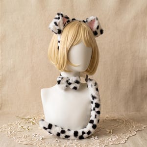 May include: A set of faux fur accessories featuring a white and black spotted dalmatian pattern. The set includes a headband with two upright cat-like ears with pink inner lining, a matching bow tie choker, and a long, plush, spotted tail.