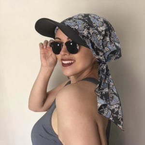 May include: A black visor with a paisley patterned head covering. The paisley pattern includes blue and white accents on a dark background. The person is wearing black sunglasses and a gray tank top.