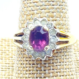 May include: A gold ring featuring a central oval-cut deep purple gemstone surrounded by a halo of small, clear, round-cut stones. The band is split into two sections on each side, leading to a polished gold shank that supports the stone setting.