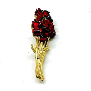 Può includere: Una spilla in metallo color oro a forma di bouquet di fiori. Il pezzo presenta petali rosso scuro lucidi sulla parte superiore, disposti a grappolo, con lunghi steli dorati testurizzati e piccole foglie che si estendono verso il basso.