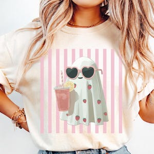 Woman wearing an ivory Comfort Colors t-shirt featuring a cute pastel ghost in heart-shaped sunglasses holding a pink drink, set against pink vertical stripes; styled with light-wash denim shorts for a trendy summerween aesthetic.
