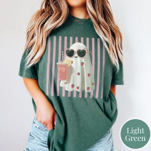 Puede incluir: Camiseta verde claro con una ilustraci&oacute;n de fantasma. El fantasma lleva gafas de sol en forma de coraz&oacute;n y est&aacute; adornado con fresas. Sostiene una bebida rosa con una rodaja de lim&oacute;n, sobre un fondo de rayas rosas. La camiseta es de corte holgado.