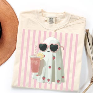 Puede incluir: Camiseta color crema con un fantasma de dibujos animados que lleva gafas de sol en forma de coraz&oacute;n y sostiene una bebida rosa con una rodaja de lim&oacute;n. El fantasma est&aacute; decorado con fresas y sobre rayas verticales rosas. La camiseta tiene una etiqueta Comfort Colors.