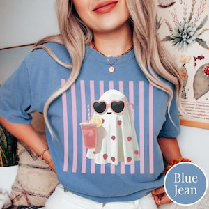 Puede incluir: Una camiseta azul vaquero con un fantasma de dibujos animados que lleva gafas de sol en forma de coraz&oacute;n y sostiene una bebida rosa con una pajita. El fantasma est&aacute; decorado con fresas y sobre un fondo de rayas rosas y blancas.
