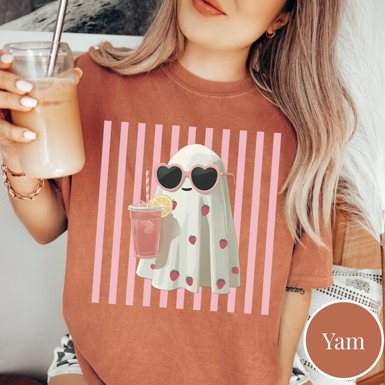 Puede incluir: Camiseta de color terracota con un fantasma de dibujos animados que lleva gafas de sol en forma de coraz&oacute;n y sostiene una bebida rosa con una pajita. El fantasma est&aacute; decorado con fresas y sobre un fondo de rayas rosas.