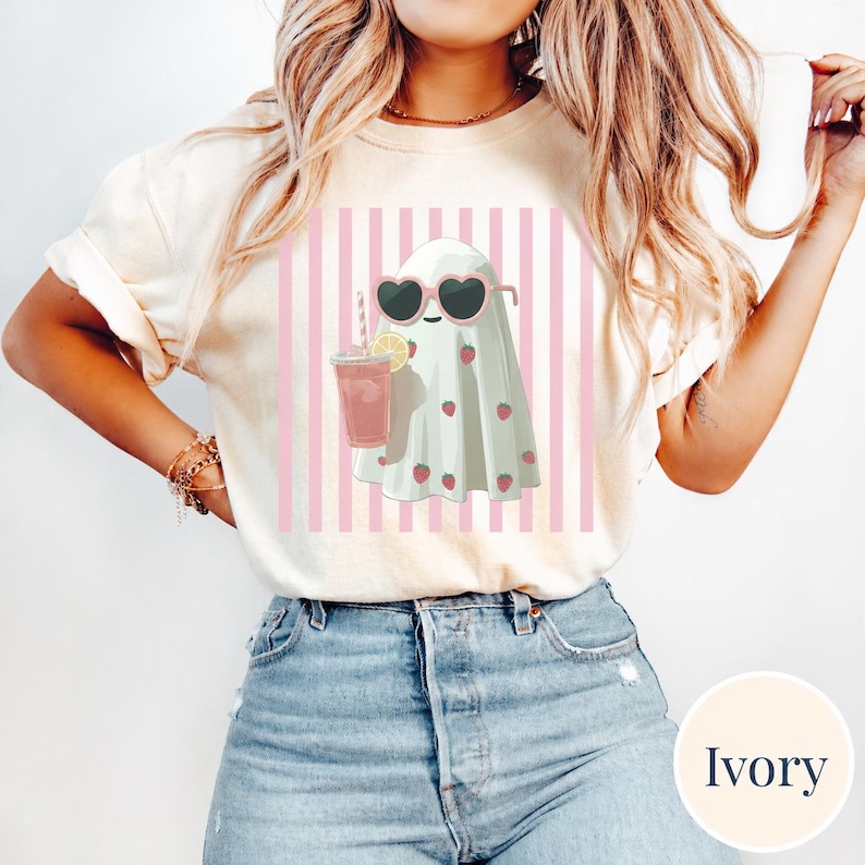 Woman wearing an ivory Comfort Colors t-shirt featuring a cute pastel ghost in heart-shaped sunglasses holding a pink drink, set against pink vertical stripes; styled with light-wash denim shorts for a trendy summerween aesthetic.