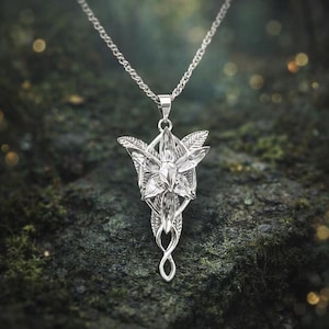 May include: A silver-colored pendant necklace featuring a stylized flower design with a central crystal and leaf accents. The pendant hangs from a delicate chain. The necklace is displayed against a textured, dark green background.