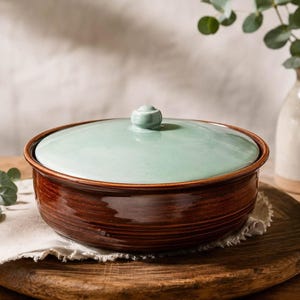 May include: A round ceramic casserole dish with a glossy dark brown base and a smooth, light mint green lid featuring a small central knob. The dish has horizontal ridged detailing along the exterior sides and a wide, flat rim.