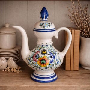 May include: A ceramic teapot with a white base, featuring a hand-painted floral design in yellow, blue, pink, and green. The teapot has a curved spout, a matching lid with a pointed finial, and blue horizontal bands around the base and neck.