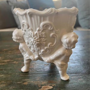 Könnte beinhalten: Ein weißer Keramik-Blumentopf mit detailliertem Design. Der Topf wird von drei Cherubfiguren getragen und weist geprägte Blumen- und Schnörkeldetails auf. Der Innenraum ist leer, und der Topf steht auf einer abgenutzten Holzoberfläche.